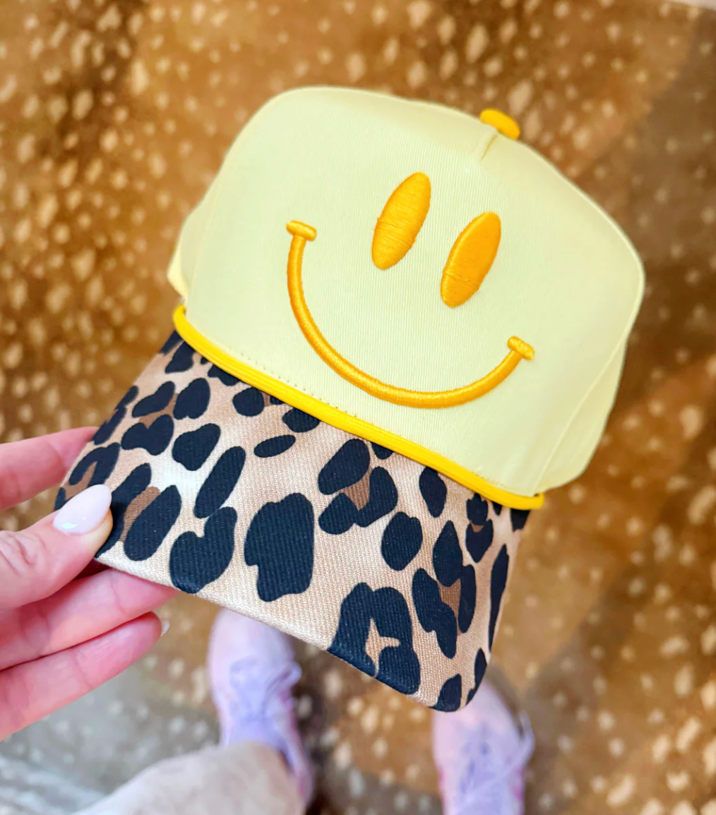 Yellow smiley face cap with leopard print brim held by a hand against a patterned background