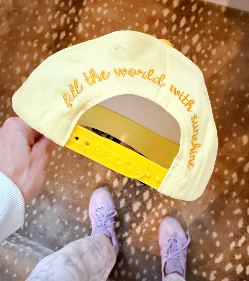 Yellow cap with 'fill the world with sunshine' text held by a person wearing pink shoes on a patterned floor.